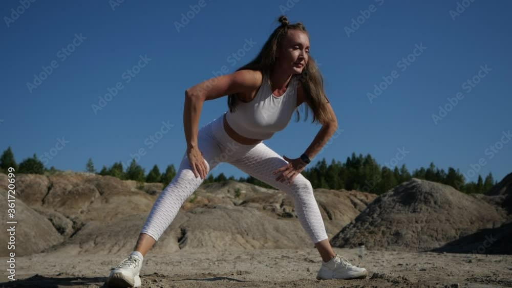 A sports girl conducts a training session in nature. Slender girl in a white suit does stretching legs. Blonde makes side lunges surrounded by sands A sports girl conducts a training session in nature. Slender girl in a white suit does stretching legs. Blonde makes side lunges surrounded by sands
