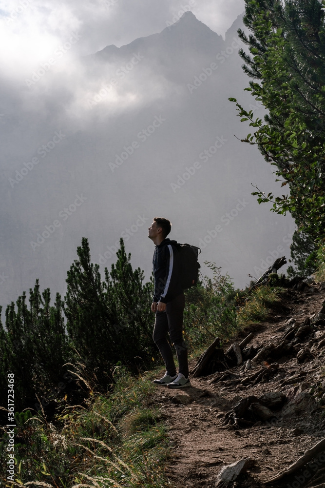 Trail to Sorapis. Dolomite Alps. Italy. Tourist man with backpack enjoy view on alpine path surrounded by greenery on background of mountains & cloud sky
