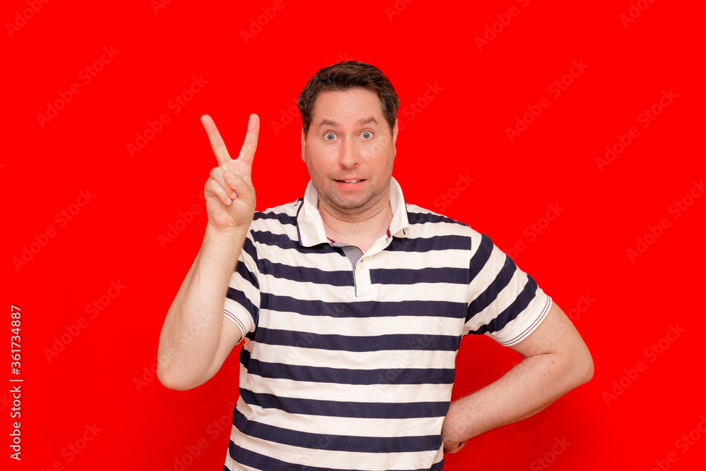 Young man wearing striped t-shirt standing over isolated red background look at camera doing victory sign.