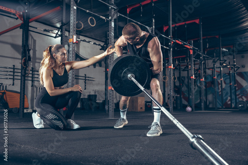 Beautiful young sporty couple training, workout in gym together. Caucasian man training with female trainer. Concept of sport, activity, healthy lifestyle, strength and power. Working out with barbell