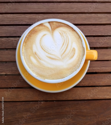 Yellow cup of coffe with milk on a dark background. Hot latte or Cappuccino decorated with foam with heart on a wooden table with copy space. Top view.