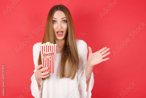 Optimistic Young caucasian woman holding bucket with popcorn raises palm from joy, happy to receive awesome present from someone, shouts loudly, Excited caucasian female screaming.