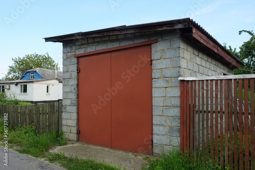 facade of a gray brick garage with red iron closed gates and a wooden fence on a rural street