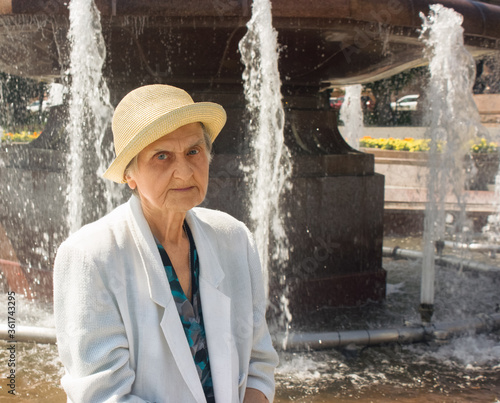 Elderly woman portrait. Senior woman in straw hat and white jacket on the background of the fontain. Social distance.