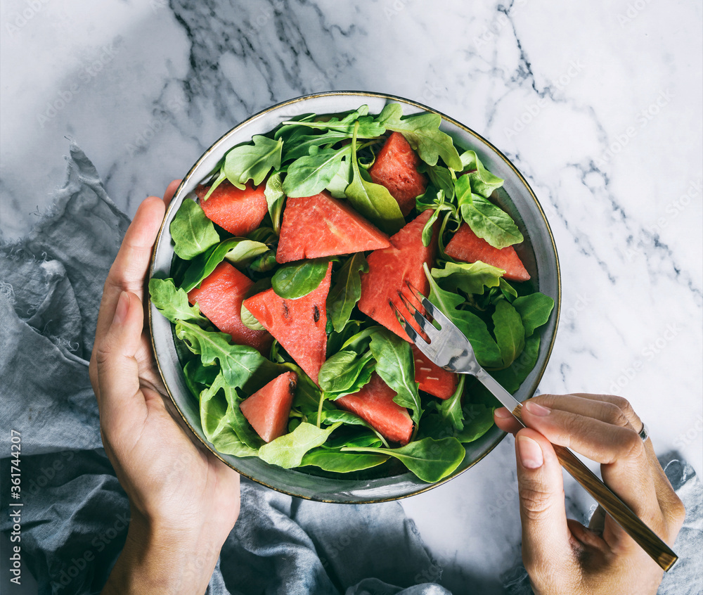 Hands holding healthy fresh summer watermelon salad with arugula, spinach and greens on light marble background. Healthy food, clean eating, Buddha bowl salad, top view