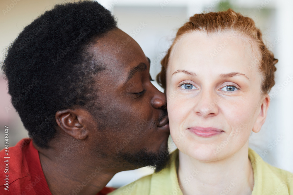 Fototapeta premium Close-up of young woman looking at camera while African man kissing her cheek