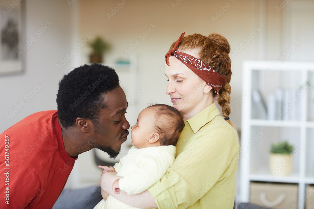 Obraz premium Young woman holding baby on her hands while African man kissing her they are at home
