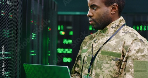 Close up of African American male army technician in camouglage costume and hat typing on keyboard of laptop computer while working with secret data at server room. Government analytic center.