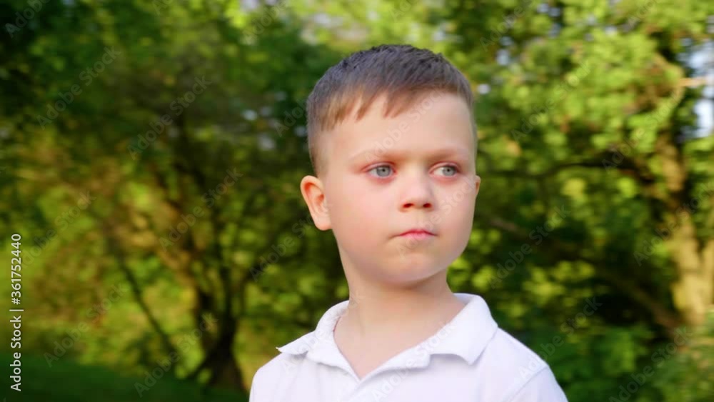 Little boy in a white T-shirt looks around in the park on a background of trees. 4K slow motion footage Little boy in a white T-shirt looks around in the park on a background of trees. 4K slow motion footage
