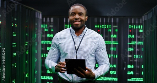 Fototapeta Portrait of young handsome African American man smiling cheerfully too camera in database center full of computers and servers