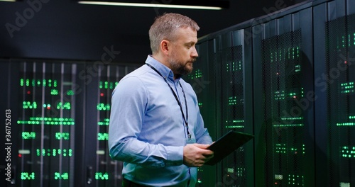Caucasian man, data base administrator in server room, tapping on tablet device and checking information on computer. Work with secret information. Big data storage. Digital defence concept.