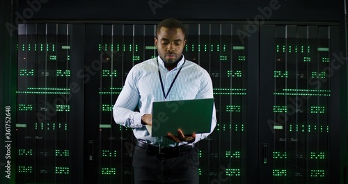 Male African American big data analytic walking at server dark room, working with secret data and using laptop computer to check the information. Monitoring database center. Cloud storage of info.