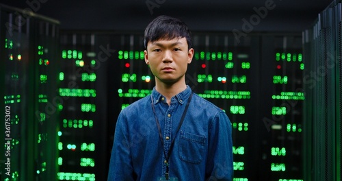 Portrait of young Asian guy looking at camera with slight smile and standing among servers in dark room. Male cybesecurity developer posing among computers.
