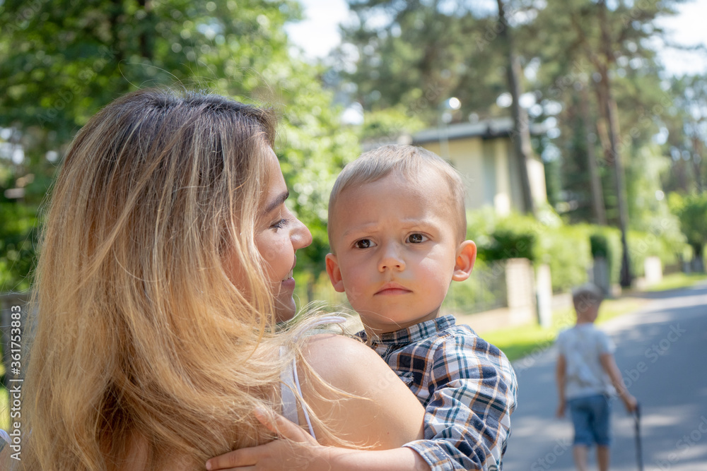 Beautiful mother holding her son in arms. Adorable mom hugging her child and looking at him while walking in the neighbourhood. Family concept.