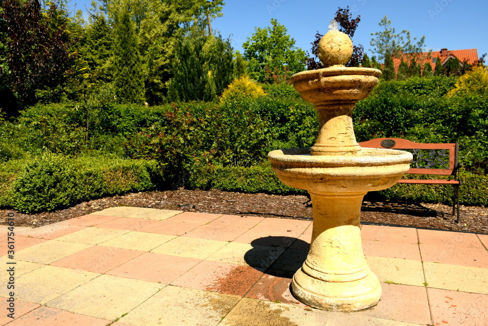 A close up on a tall clay ecru fountain or well standing in the middle of a decorative, well maintained garden next to a wooden and metal bench seen on a cloudless summer sky in Poland