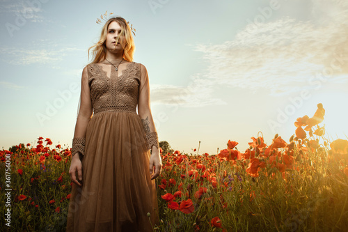 A beautiful girl is standing in a fashionable dress and a wreath in a poppy field. Fashion. Lifestyle.