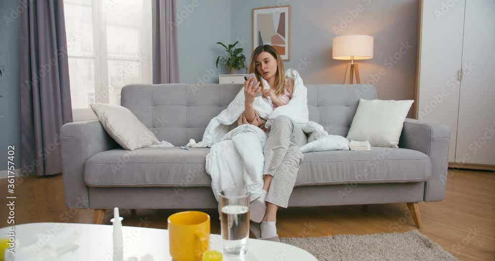 Unhealthy young Caucasian woman with fever sitting on couch in living room and videochatting on smartphone. Sick girl caughing and having videochat on mobile phone via webcam. Unhealthy young Caucasian woman with fever sitting on couch in living room and videochatting on smartphone. Sick girl caughing and having videochat on mobile phone via webcam.