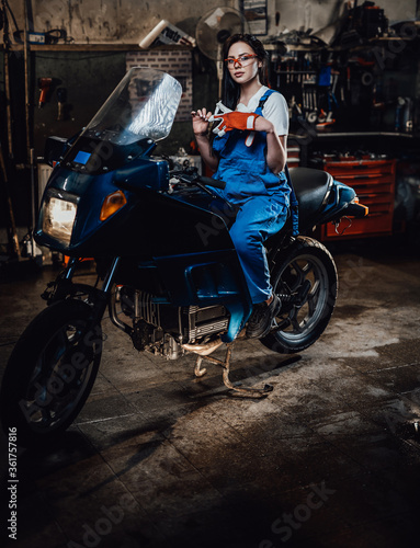 Beautiful young brunette female mechanic in blue overalls sitting on sportbike in garage or workshop
