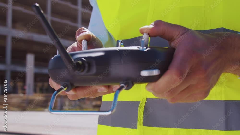 Drone operator holding remote controller. Professional builder in helmet and vest standing in front of construction site. Office building and crane background. Business, real estate and investment. Drone operator holding remote controller. Professional builder in helmet and vest standing in front of construction site. Office building and crane background. Business, real estate and investment.