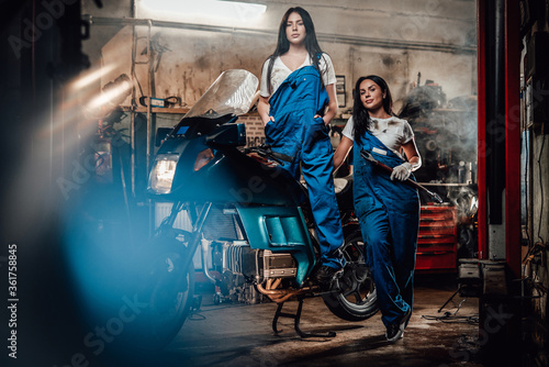 Two hot brunette women in blue overalls posing next to a sportbike in authentic workshop garage