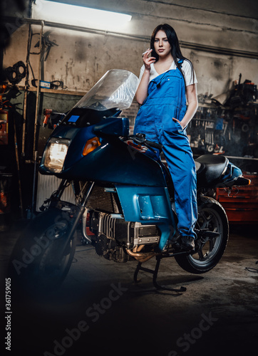 Young female mechanic relaxing smoking a cigarette while standing on sportbike in garage or workshop