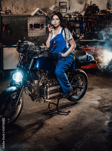 Beautiful brunette female mechanic in blue overalls relaxing smoking a cigarette while sitting on custom bobber in garage or workshop © Fxquadro
