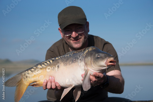Happy smiling fisherman holding his fish with lake on the background. Selective focus on the carp.