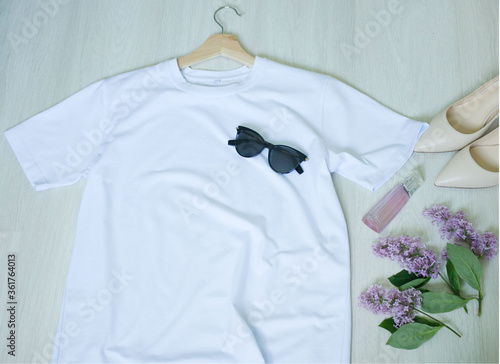 white t-shirt on a hanger and accessories, black glasses, a bottle of perfume, a sprig of lilac and shoes, top view. Fashion, style, summer look, wardrobe