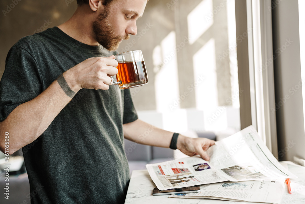 Photo of serious redhead man reading newspaper while drinking tea Photo of serious redhead man reading newspaper while drinking tea
