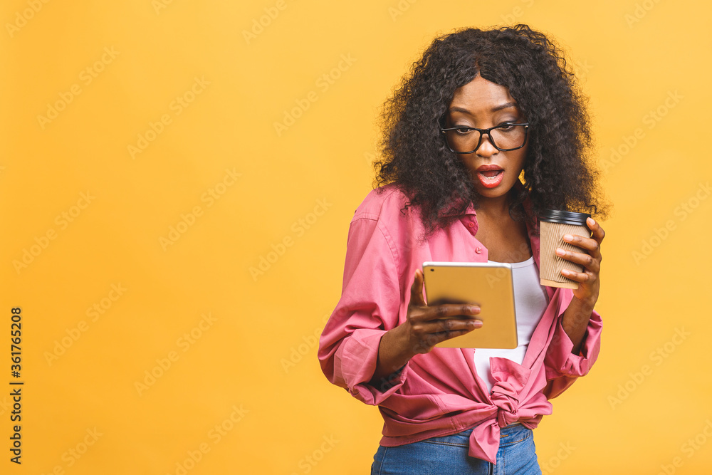 Portrait of african american woman 20s with afro hairdo looking aside while drinking takeaway coffee or tea from paper cup isolated over yellow background. Using tablet. Portrait of african american woman 20s with afro hairdo looking aside while drinking takeaway coffee or tea from paper cup isolated over yellow background. Using tablet.