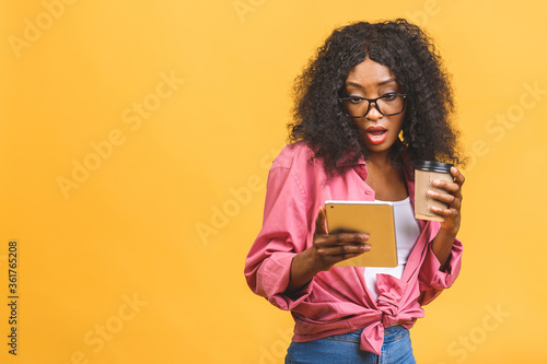 Portrait of african american woman 20s with afro hairdo looking aside while drinking takeaway coffee or tea from paper cup isolated over yellow background. Using tablet.