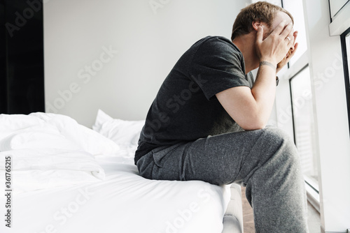 Photo of displeased redhead man covering his face while sitting on bed