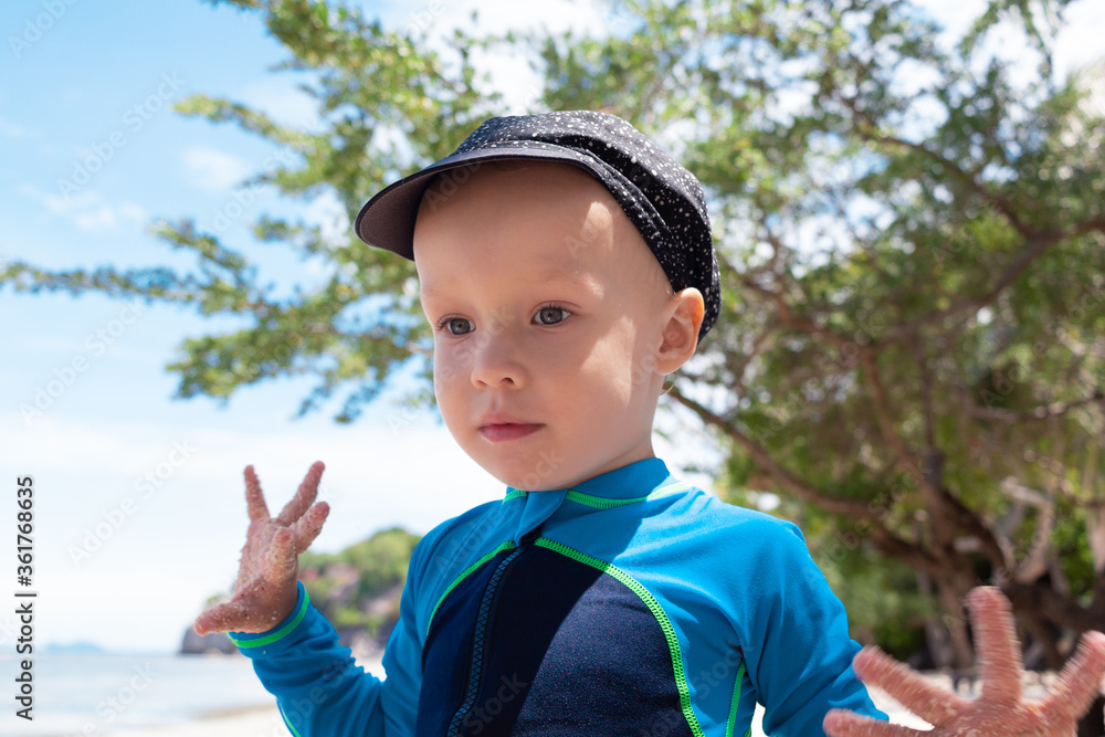 Young boy in blue swimsuite and black cap playing with sand on a tropical beach with a sea, sky and green tree back ground. Summer time vacation consept. Young boy in blue swimsuite and black cap playing with sand on a tropical beach with a sea, sky and green tree back ground. Summer time vacation consept.