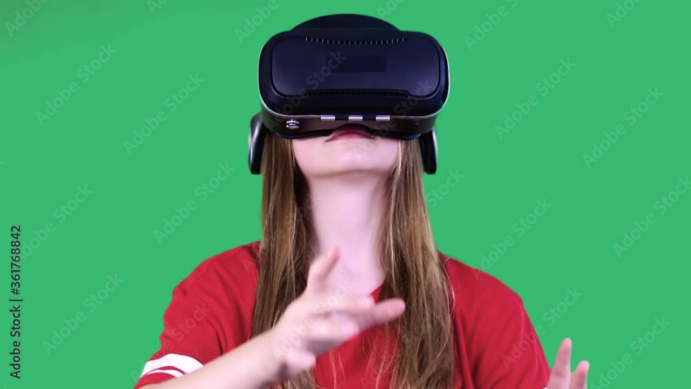 Happy excited young girl with virtual reality headset or 3d glasses, trying to touch the objects and to find the exit, on green background at studio. Modern technology concept Happy excited young girl with virtual reality headset or 3d glasses, trying to touch the objects and to find the exit, on green background at studio. Modern technology concept