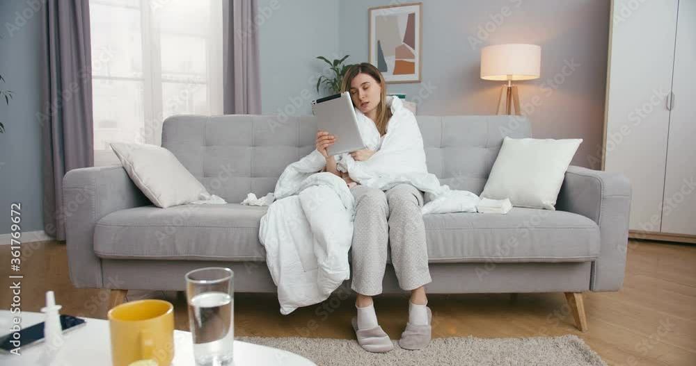 Unhealthy young Caucasian woman with fever sitting on couch in living room and videochatting on tablet. Sick girl caughing and having videochat on computer via webcam Female talking in video on device Unhealthy young Caucasian woman with fever sitting on couch in living room and videochatting on tablet. Sick girl caughing and having videochat on computer via webcam Female talking in video on device