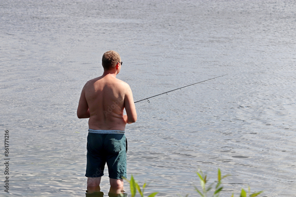 Fisherman in shorts standing in shallow water with a fishing rod, rear view. Man angling in summer on the river coast
