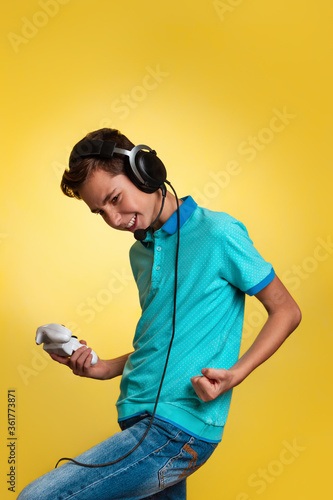 The concept of virtual and computer games. A teenage boy in a blue t-shirt and headphones, with a joystick in his hands, rejoices in winning the game. Yellow background. Copy space. Vertical © _KUBE_