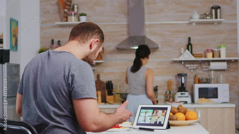 Freelancer during a video conference on tablet computer while the wife is cooking breakfast in kitchen. Entrepreneur enjoying a cup of of coffee during a video conference with coworkers. Freelancer during a video conference on tablet computer while the wife is cooking breakfast in kitchen. Entrepreneur enjoying a cup of of coffee during a video conference with coworkers.