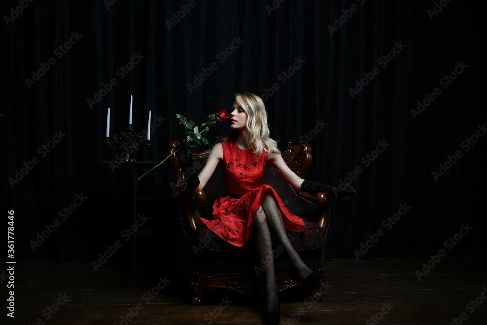 Fototapeta premium beautiful girl in a red dress posing in a chair with a red rose on a black background