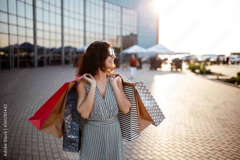 Young beautiful happy woman walks out of shopping mall with purchases. Shopaholic girl glad to buy clothes and stands outdoor with bags and credit card. Shop, sales, discount, spend money concept.