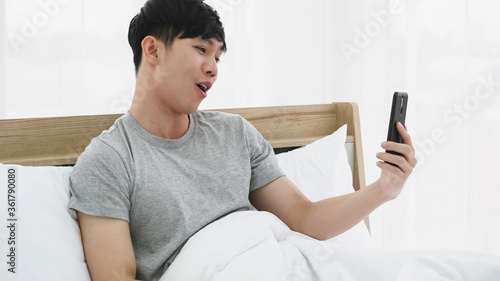 Young happy man using mobile phone for video call in bed at home