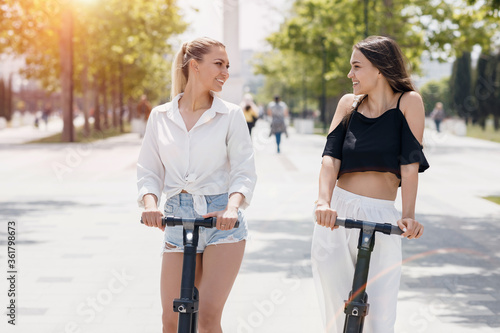 Two women friends riding electric scooters in park