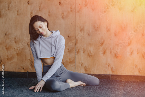 Young attractive fitness girl sitting on the floor near the window on the background of a wooden wall, resting on yoga classes