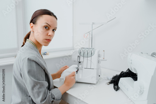 Female tailor with a centimeter ribbon over the neck is adjusting the overlock machine/ serger on the table in her mini tailoring workshop. photo