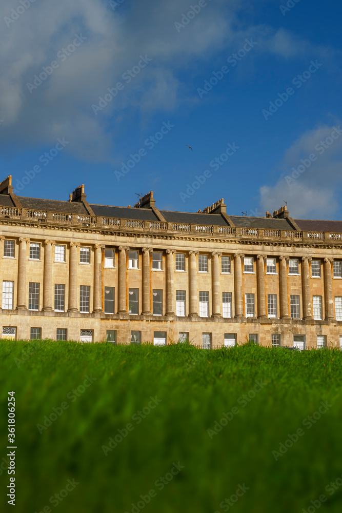 Low angle view of the Royal Crescent in Bath, United Kingdom with lush green lawn in the foreground and blue summer sky in the background.