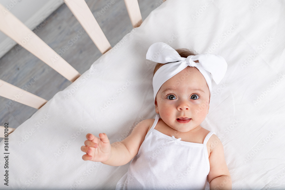 portrait of a smiling baby girl 6 months old lying in a crib in the children's room on her back with a bow on her head, looking at the camera, baby's morning, children's goods concept portrait of a smiling baby girl 6 months old lying in a crib in the children's room on her back with a bow on her head, looking at the camera, baby's morning, children's goods concept
