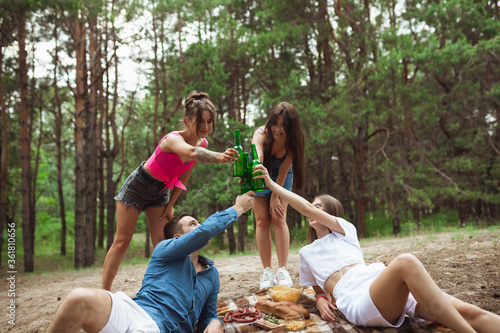 Togetherness. Group of friends clinking beer bottles during picnic in summer forest. Lifestyle, friendship, having fun, weekend and resting concept. Looks cheerful, happy, celebrating, festive.