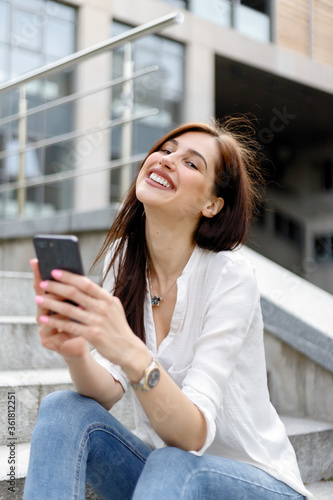 Attractive young woman in the city. Caucasian businesswoman using smartphone with hand. Portrait stylish business woman in fashionable clothes holding Phone near office building.