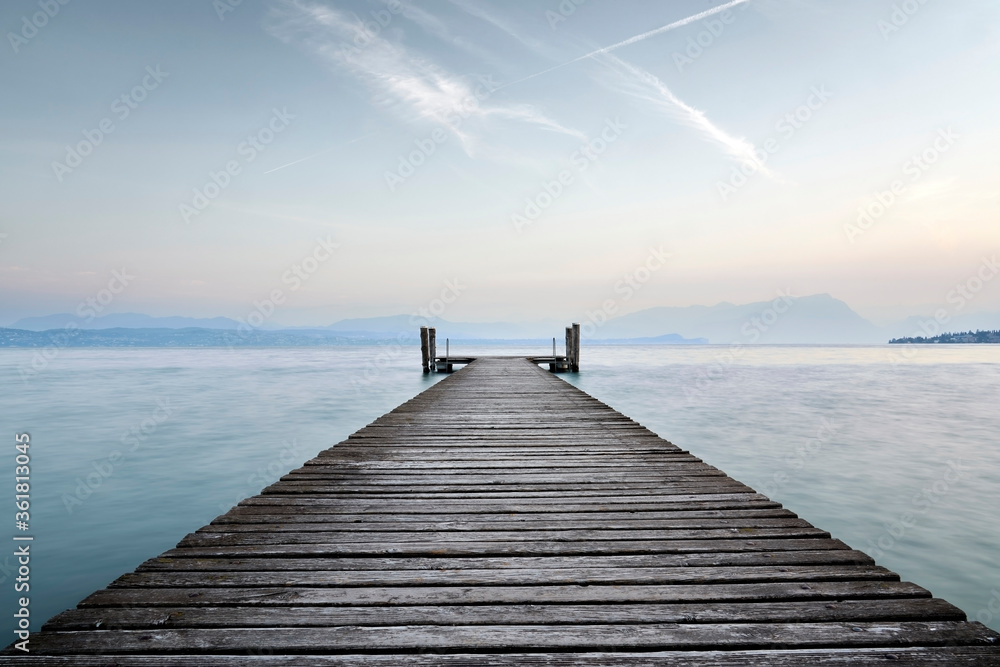 Obraz premium An old wooden pier extends into the clear blue waters of the lake. A solitary path towards the calm and peaceful silence of nature, with the sweet sound of the waves.