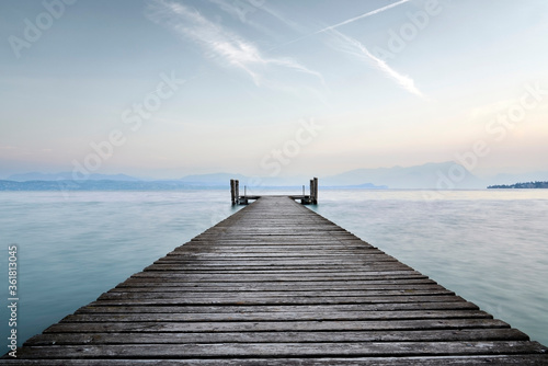 An old wooden pier extends into the clear blue waters of the lake. A solitary path towards the calm and peaceful silence of nature, with the sweet sound of the waves.